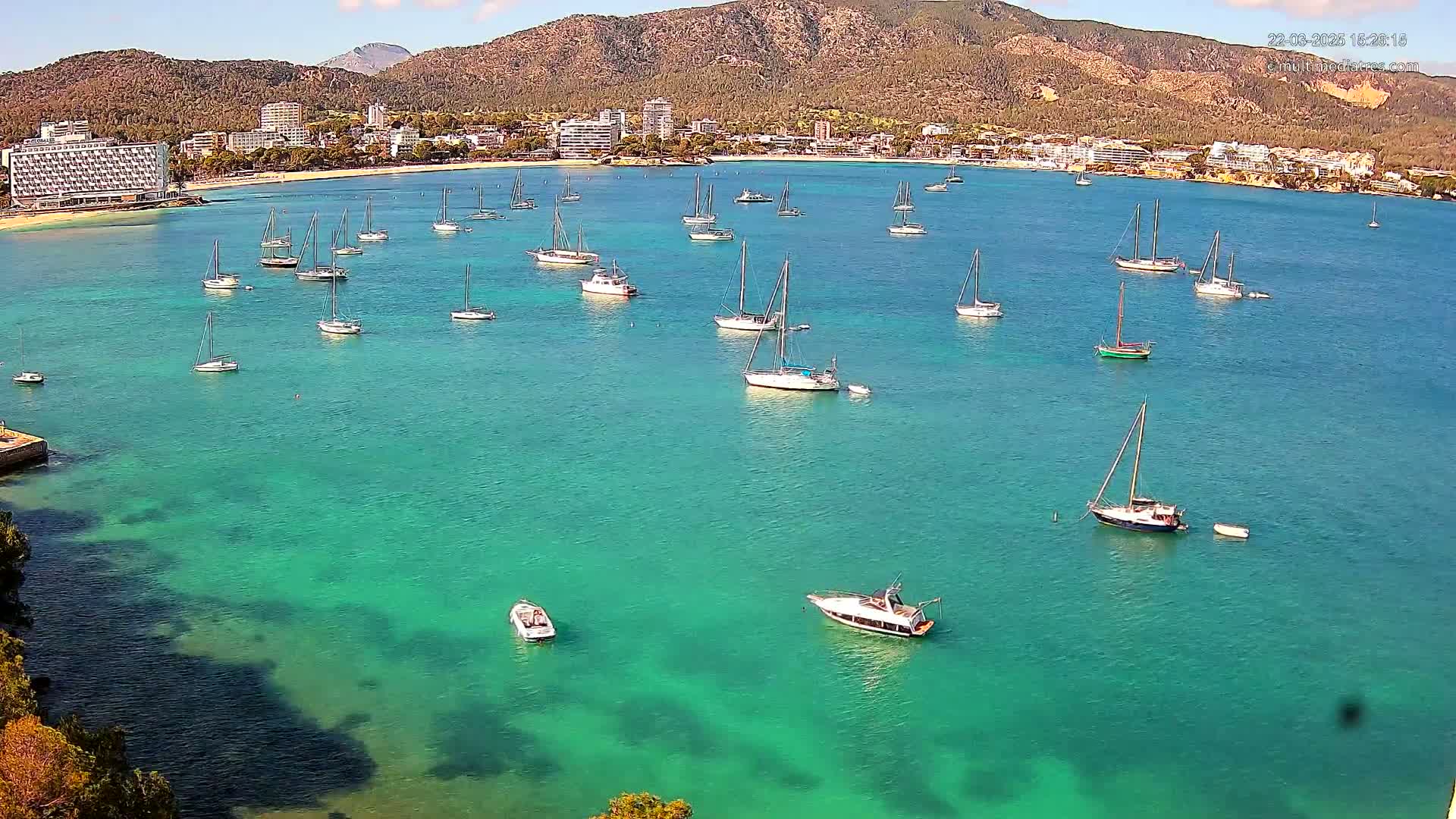 Numerous sailboats are anchored in a calm, teal-colored bay, with a beach and buildings visible in the background under a clear sky.