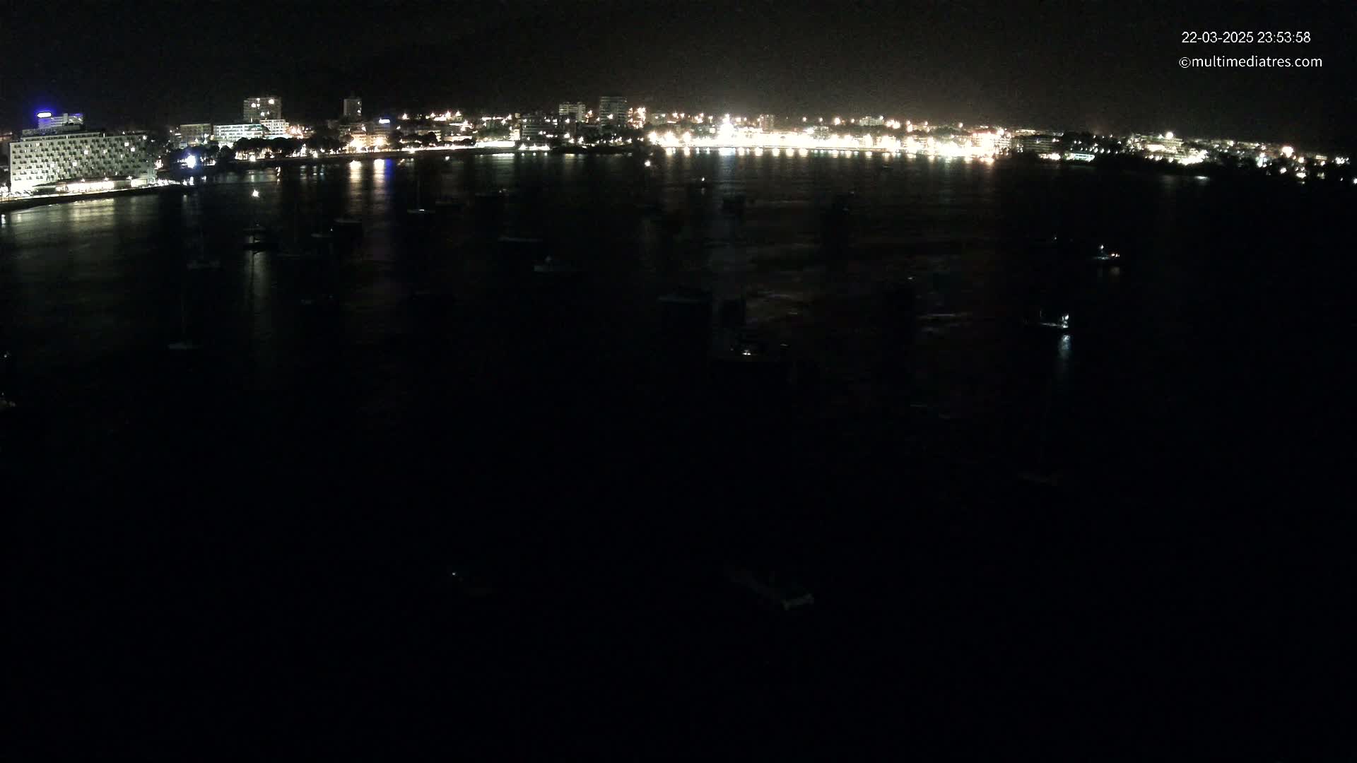 A nighttime, dark view of a city's waterfront illuminated with lights, showing numerous small boats at anchor in calm water.