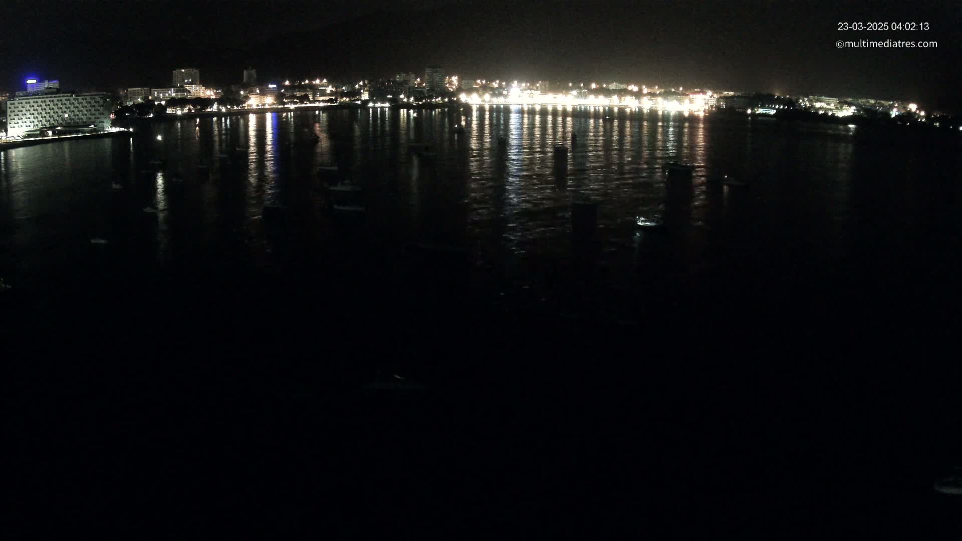At night, a city's lights are reflected on a dark body of water where several boats are moored.