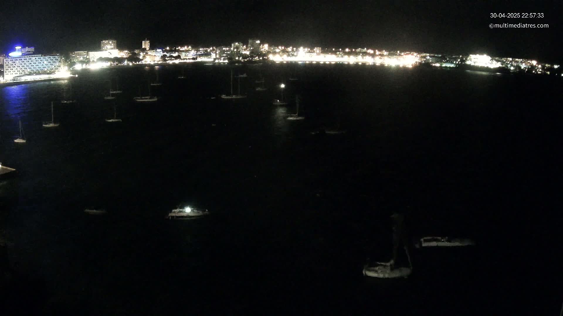 At night, a dark body of water dotted with numerous small boats is seen in front of a brightly lit city shoreline.