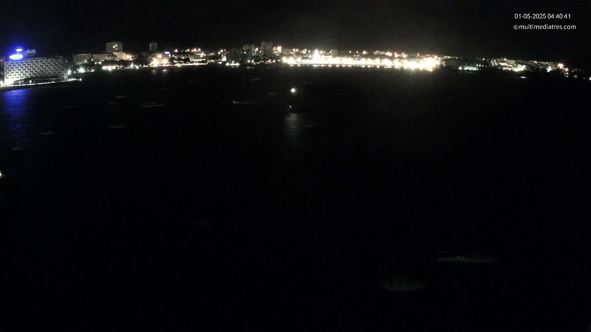 A nighttime, long shot shows a dark body of water with sparsely scattered lights, in front of a brightly lit coastline with buildings.