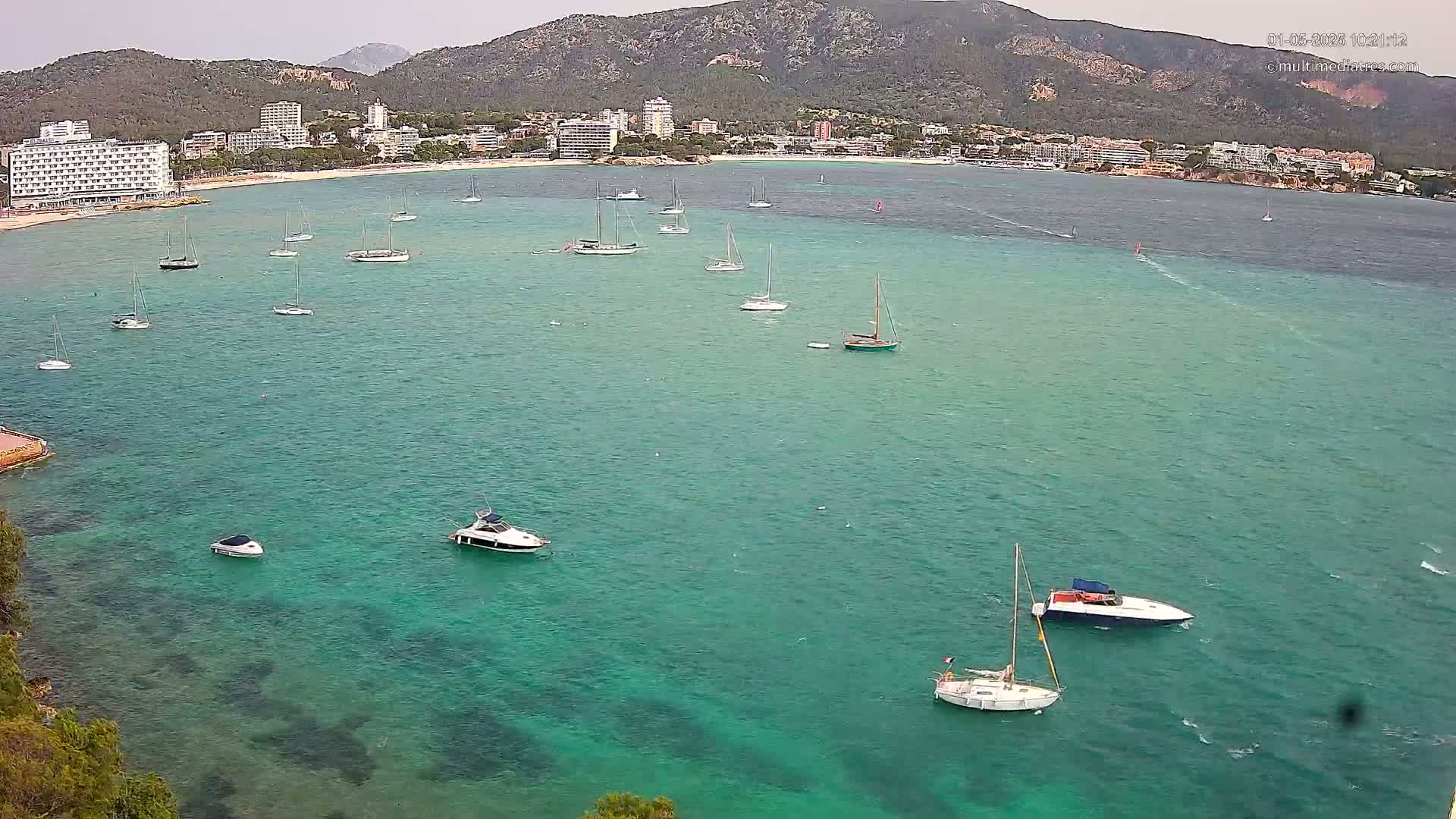 An aerial view shows numerous sailboats and motorboats in a teal bay, with a beach and buildings along a mountainous coastline under an overcast sky.