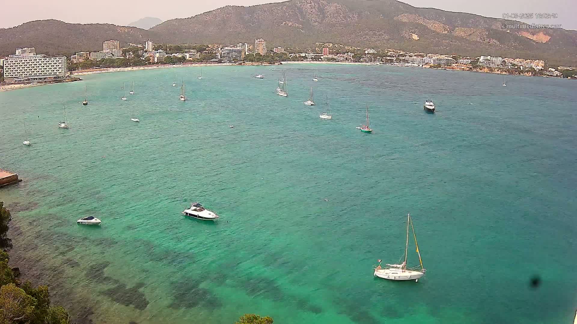 A wide shot shows a calm, turquoise bay with numerous sailboats and motorboats anchored near a beach and hotels nestled against a mountainous backdrop under an overcast sky.