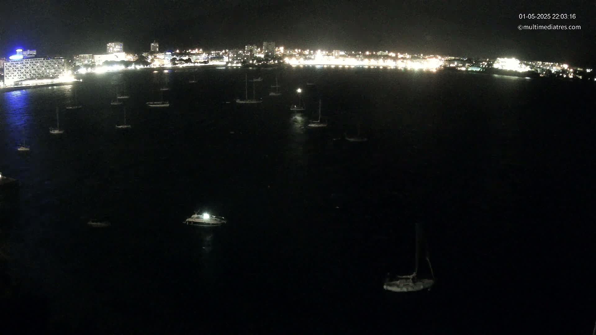 At night, numerous boats are moored in a dark body of water in front of a brightly lit city skyline.