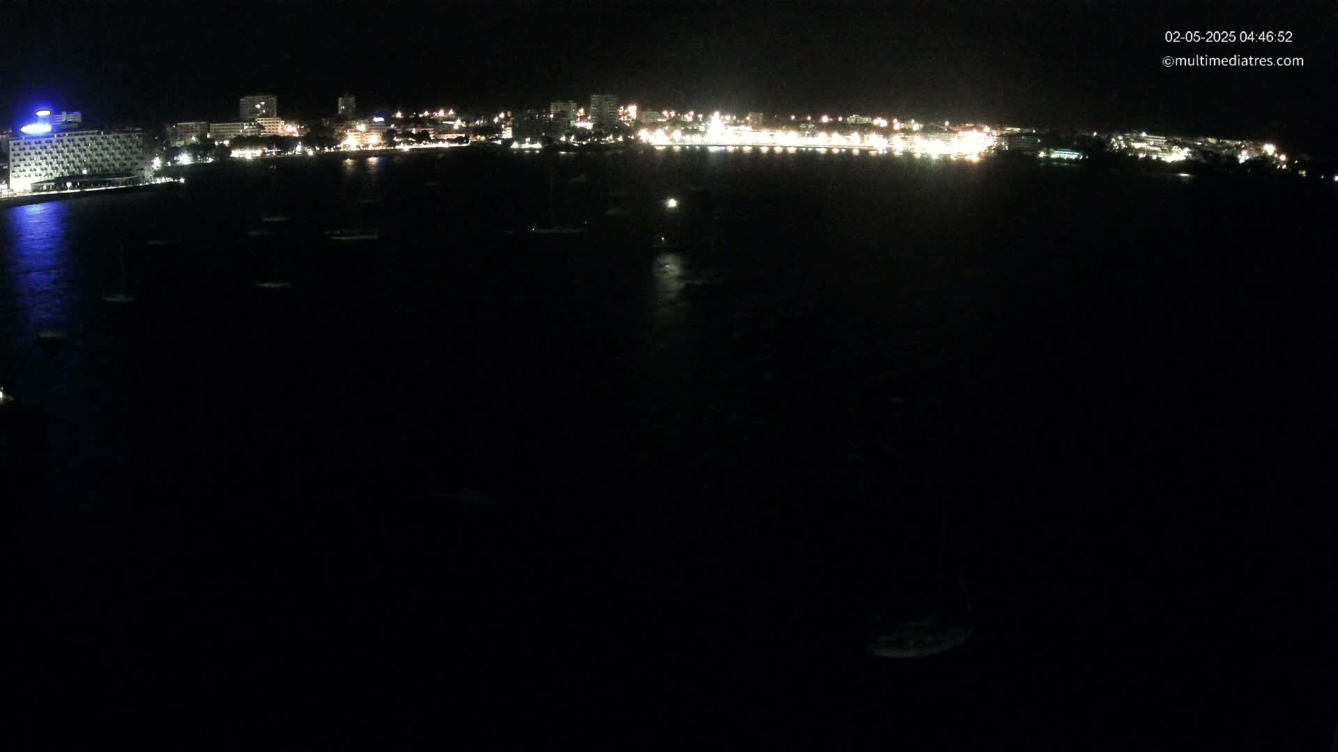 At night, a dark body of water with several boats is in front of a city's brightly lit shoreline.