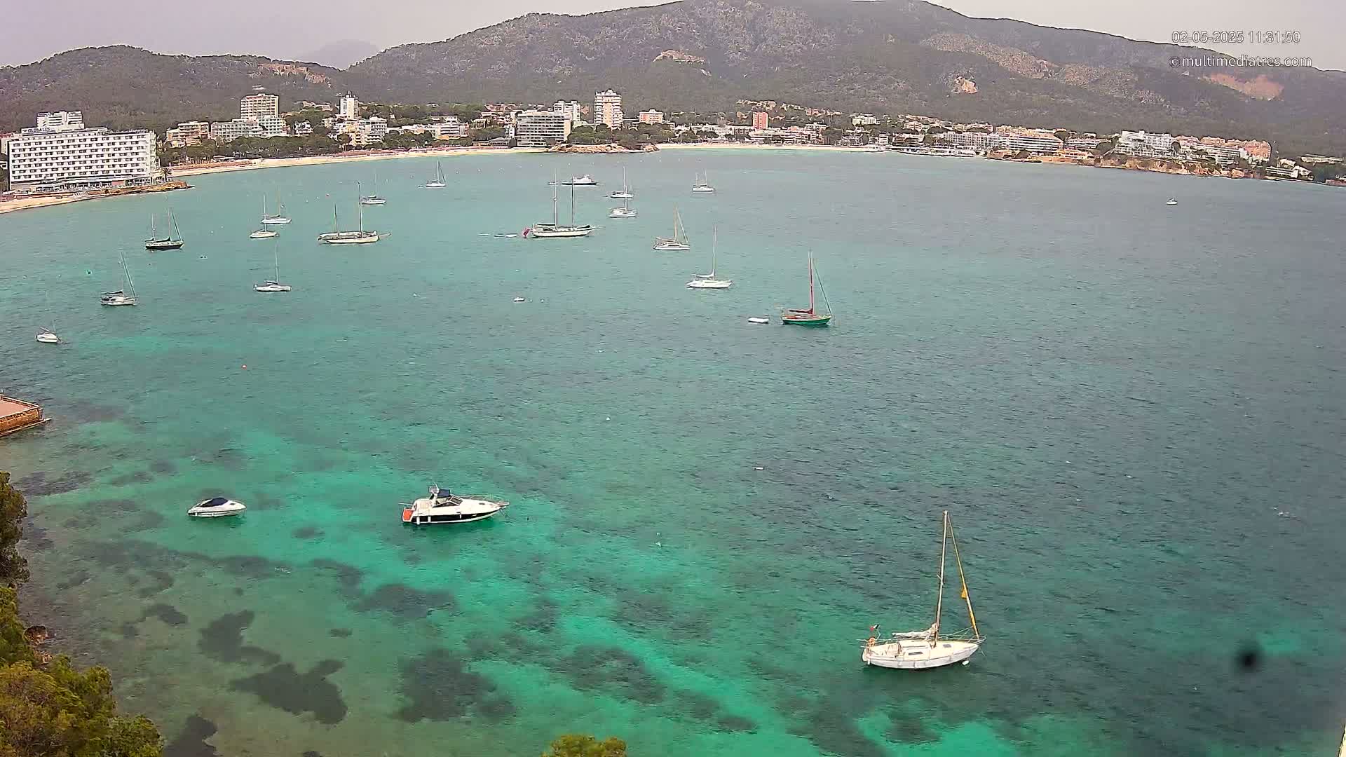 A calm, teal-colored bay with numerous sailboats and motorboats is nestled between a sandy beach and a mountainous coastline under an overcast sky.
