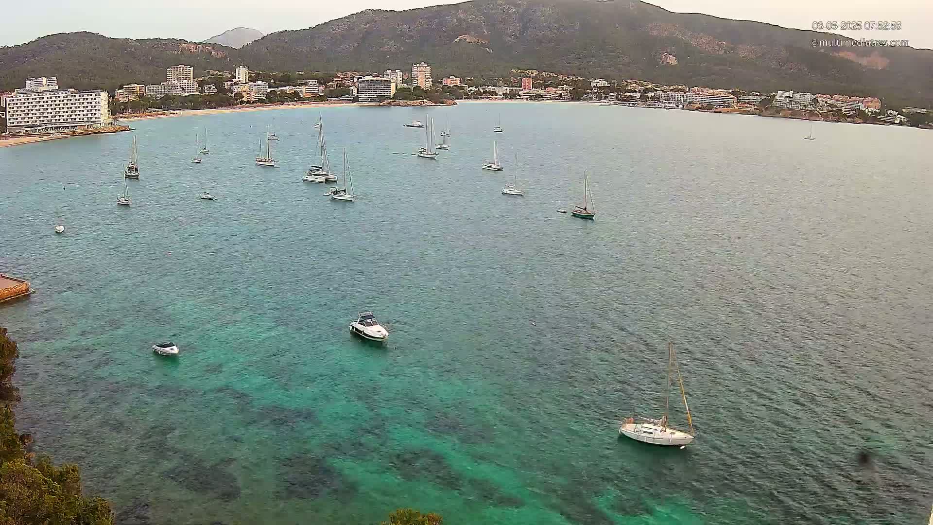 A calm bay with numerous sailboats and motorboats is seen from an elevated viewpoint, with a coastline featuring hotels and green hills in the background under an overcast sky.
