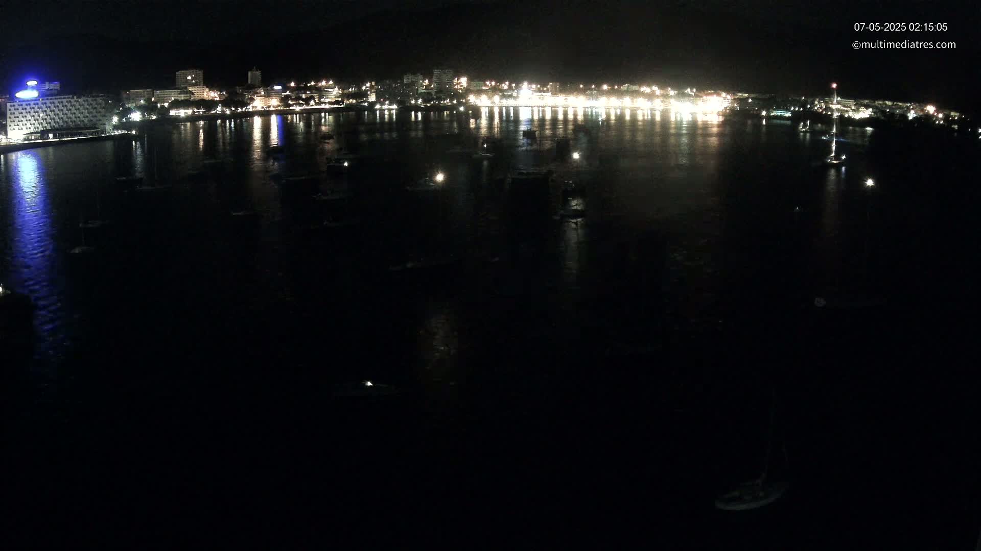At night, a dark body of water reflects the lights of a city skyline, with numerous small boats scattered across its surface.