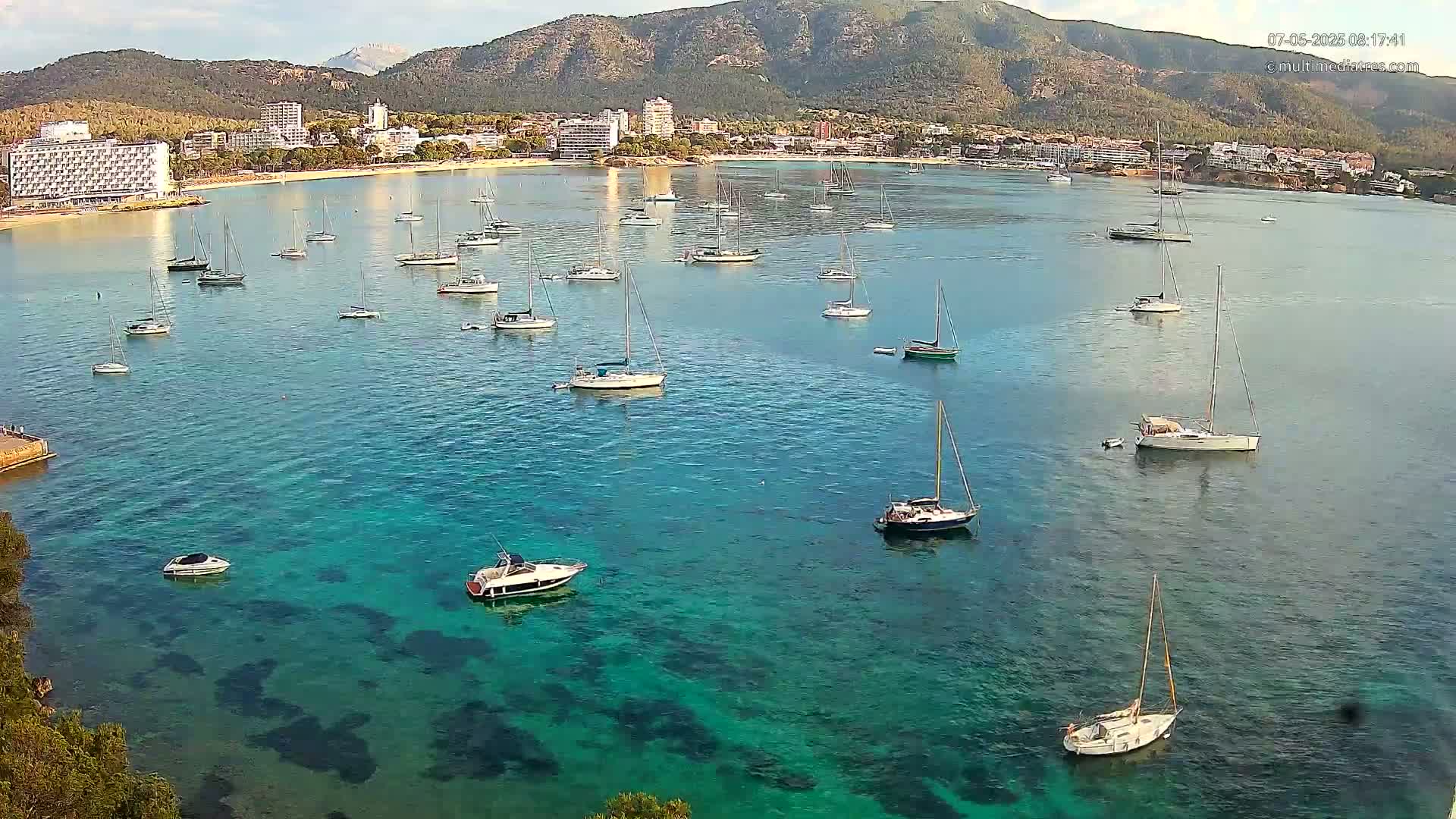 Numerous sailboats and motorboats are anchored in a calm, clear, teal-colored bay, with a coastline featuring hotels and mountains in the background.