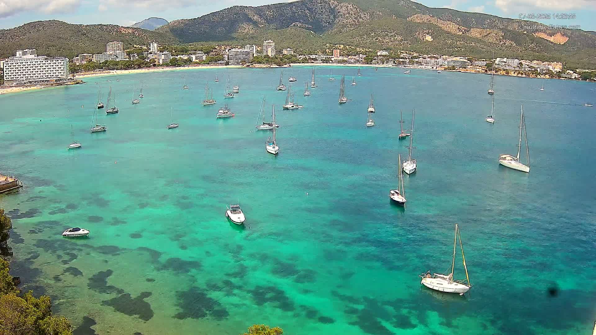 Numerous sailboats and motorboats are anchored in a calm, clear, teal-colored bay, with a mountainous coastline and hotels visible in the background under a partly cloudy sky.
