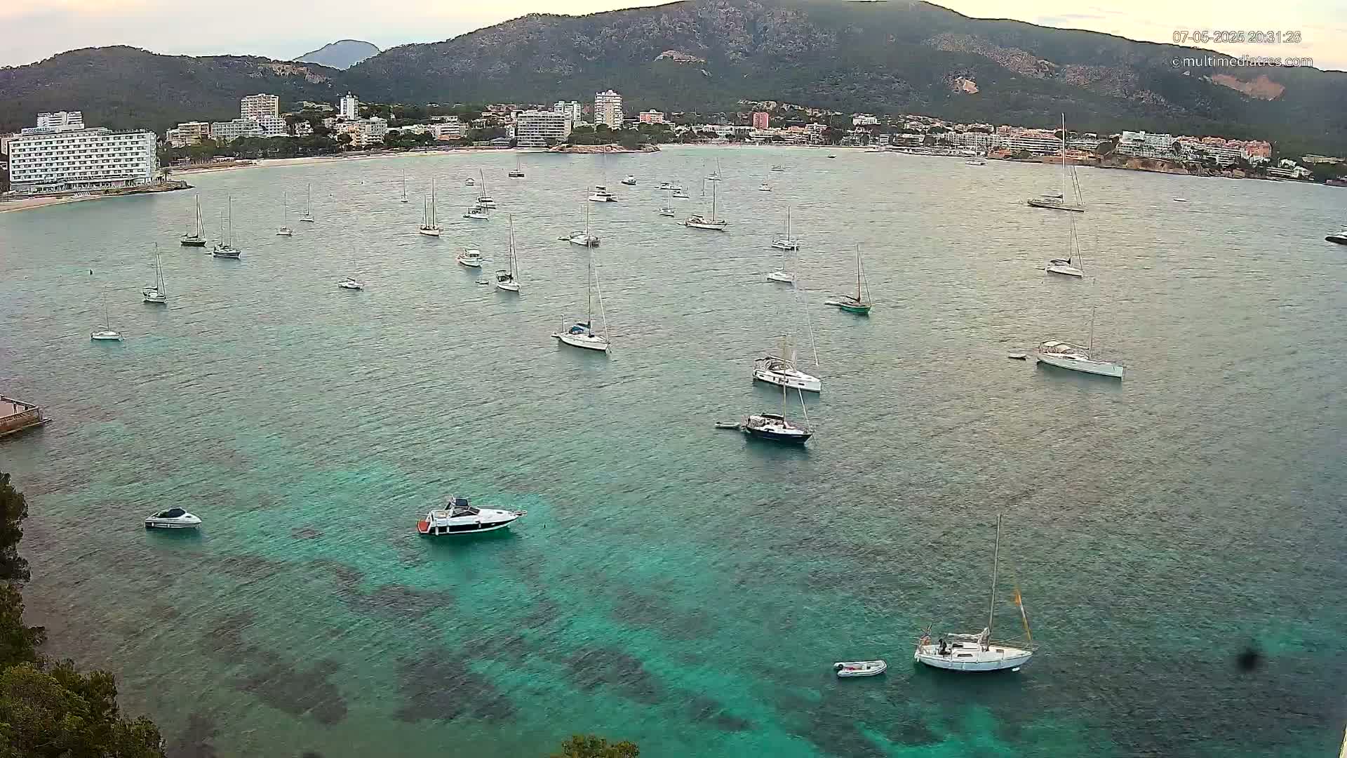 Numerous sailboats and motorboats are anchored in a calm, teal-colored bay, with a mountainous coastline and buildings in the background.