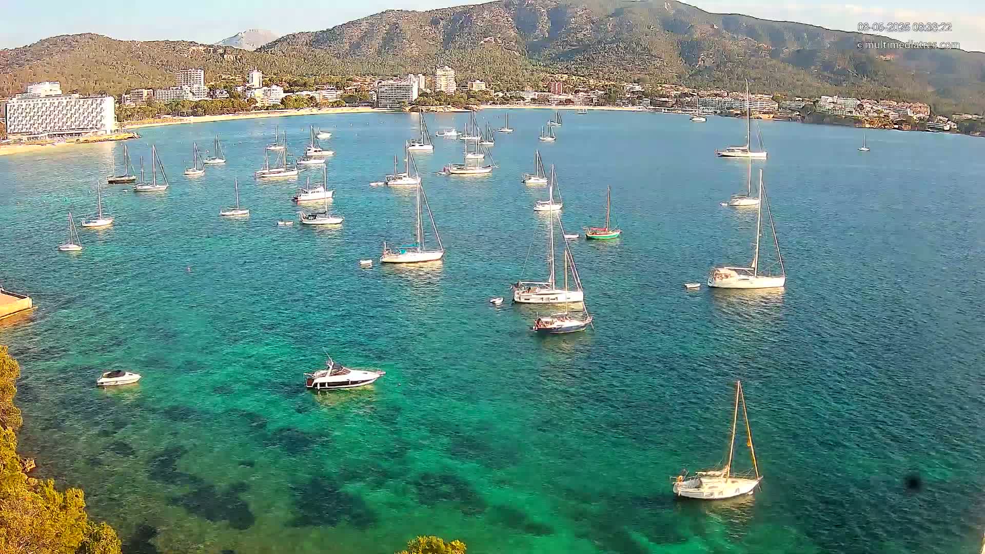 Numerous sailboats and motorboats are anchored in a calm, clear, turquoise bay near a coastline with hotels and a mountain in the background under a sunny sky.