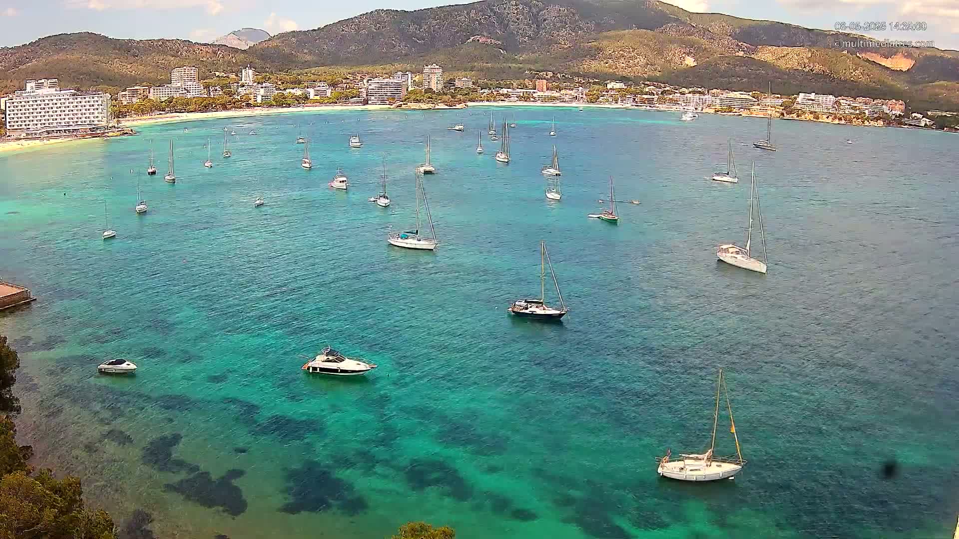 Numerous sailboats and motorboats are anchored in a calm, clear turquoise bay, with a mountainous coastline and buildings in the background under a partly cloudy sky.