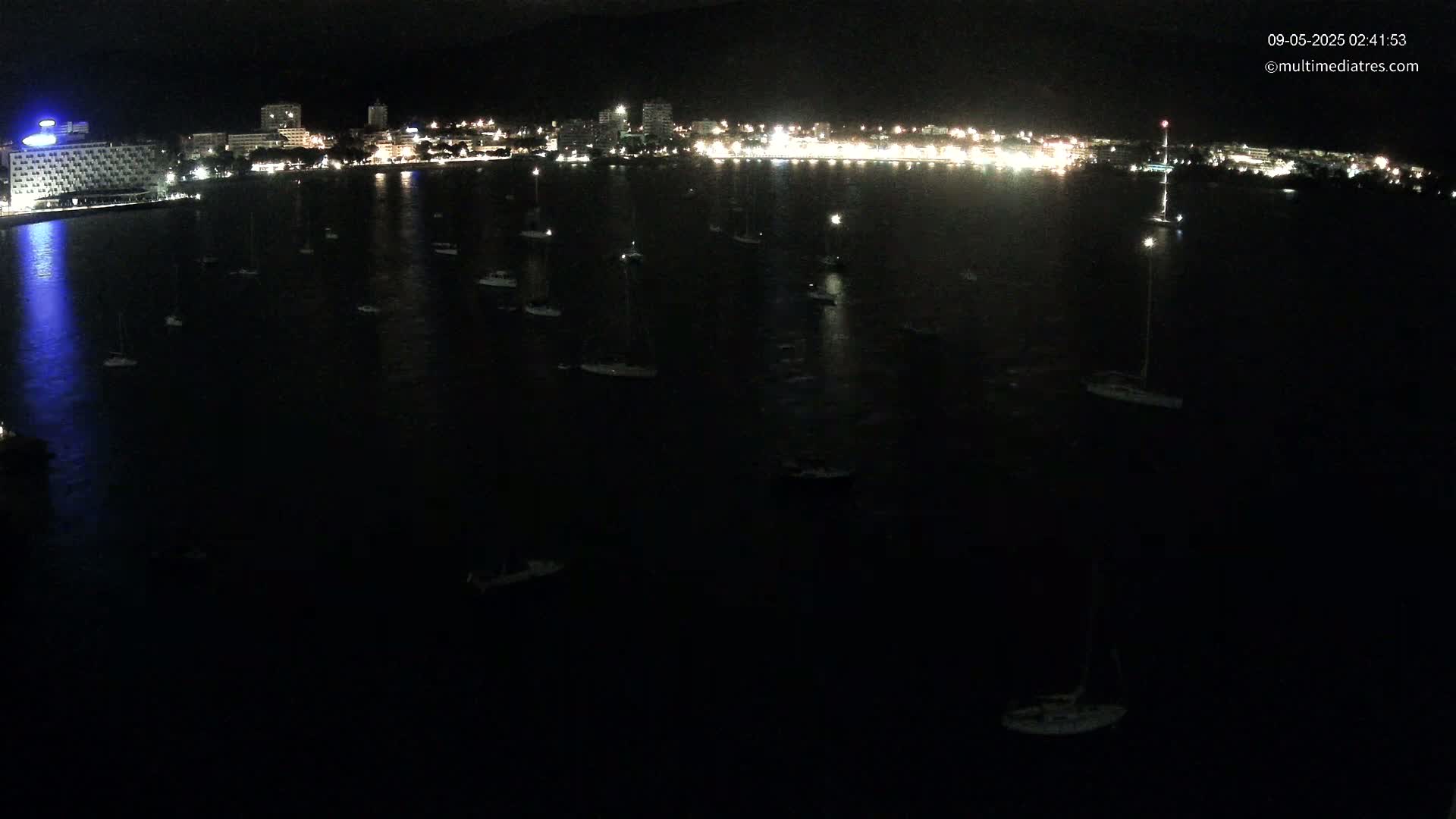 At night, a dark body of water dotted with numerous small boats is seen before a brightly lit shoreline.