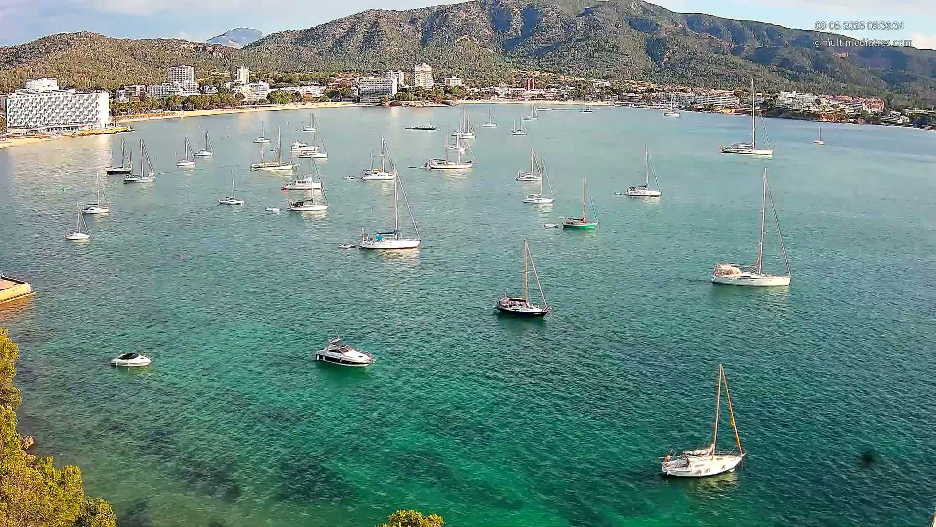 Numerous sailboats and motorboats are anchored in a calm, clear turquoise bay near a mountainous coastline with hotels and buildings.