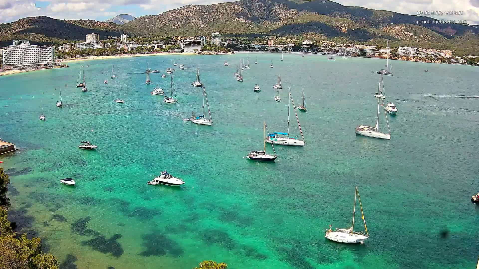 A calm, bright, turquoise bay with numerous sailboats and motorboats anchored near a shoreline with hotels and mountains in the background.