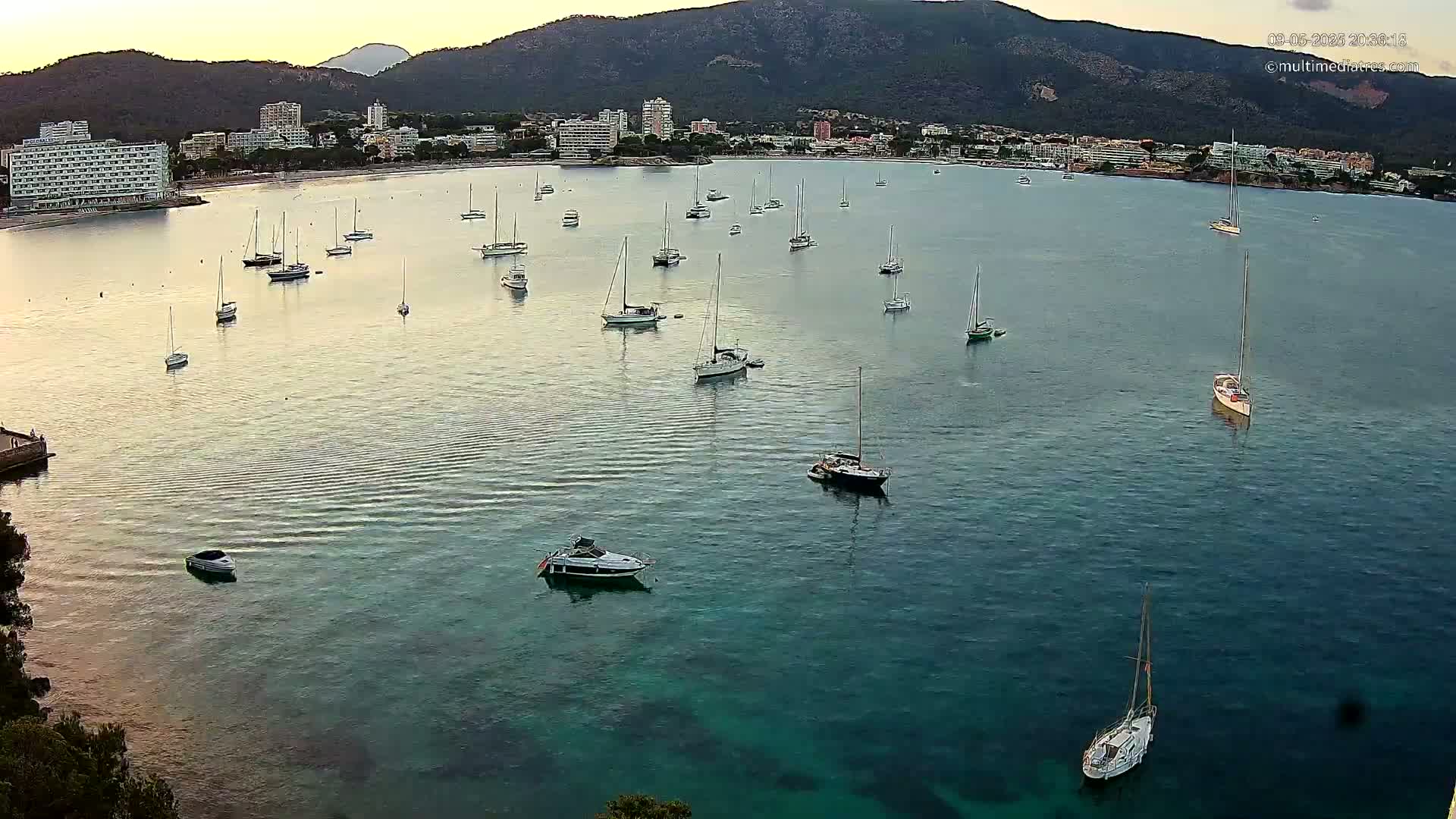 Numerous sailboats and motorboats are anchored in a calm, clear bay near a coastline with hotels and buildings nestled against a mountain under a twilight sky.