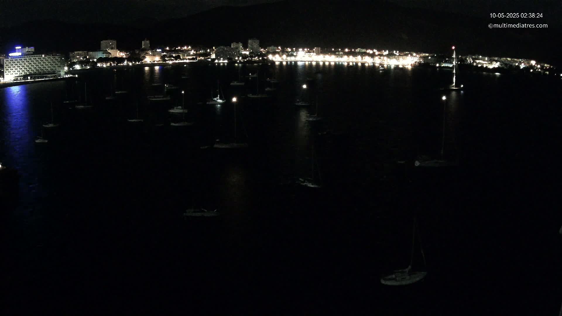 Many boats are moored in a dark, calm body of water at night in front of a brightly lit city.