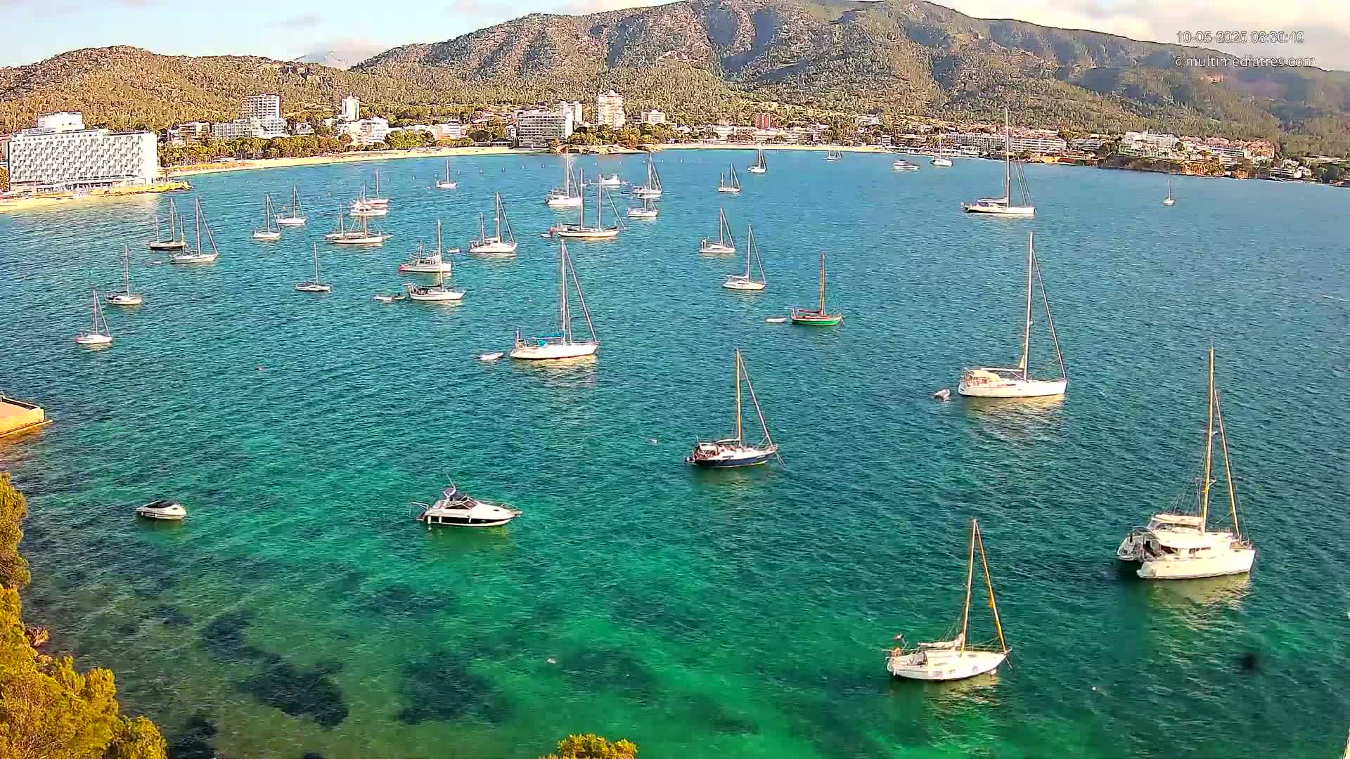 Numerous sailboats and motorboats are anchored in a calm, clear turquoise bay, with a coastline of hotels and mountains visible in the background.