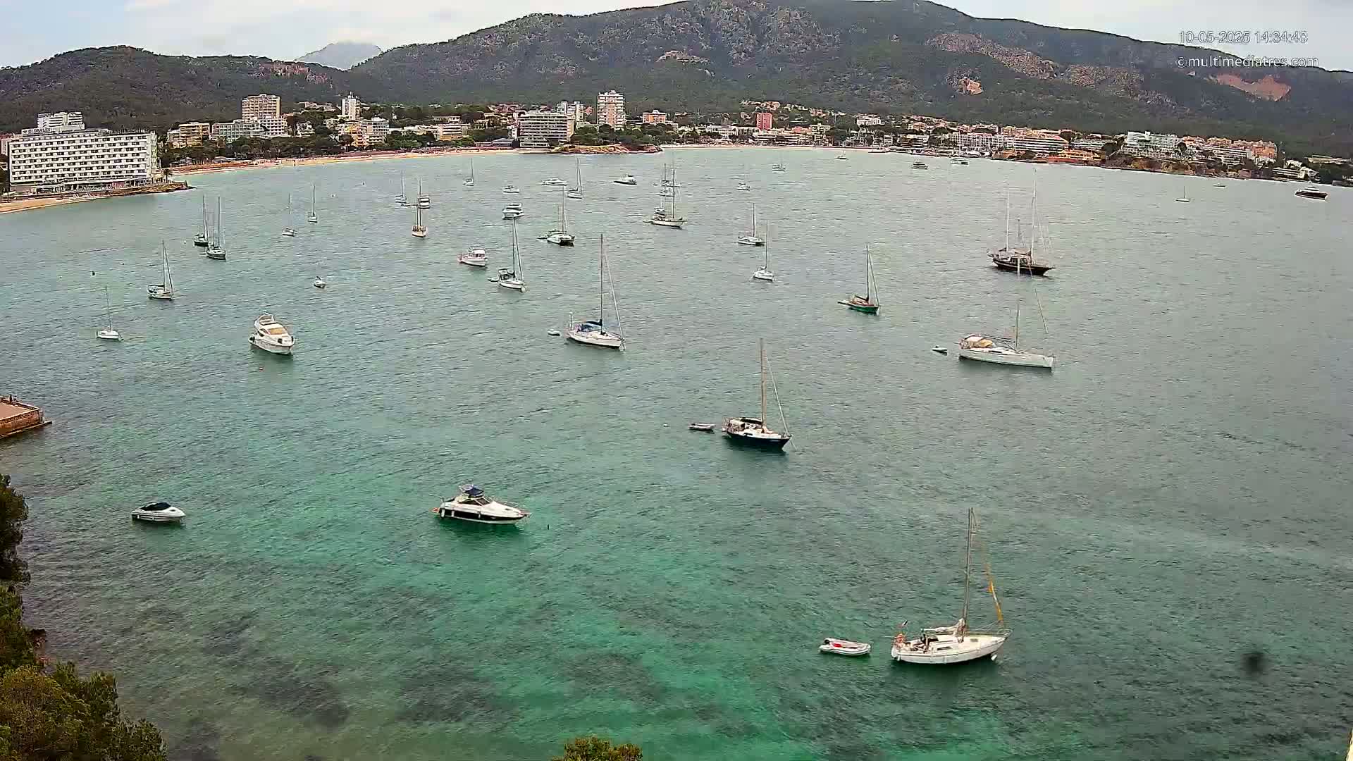 Numerous sailboats and motorboats are anchored in a calm, teal-colored bay near a mountainous coastline under a partly cloudy sky.