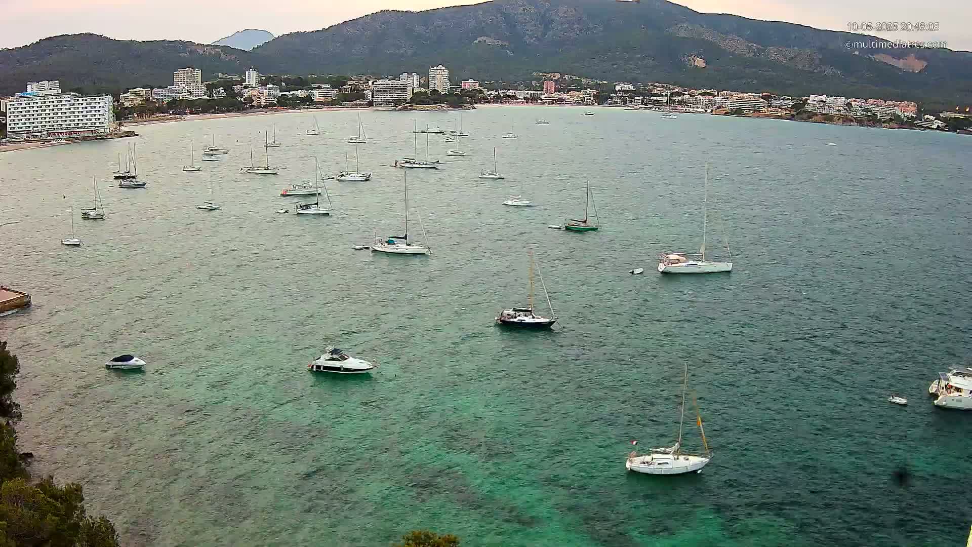 Numerous sailboats and motorboats are anchored in a calm, teal-colored bay, with a town and mountains visible in the background under an overcast sky.