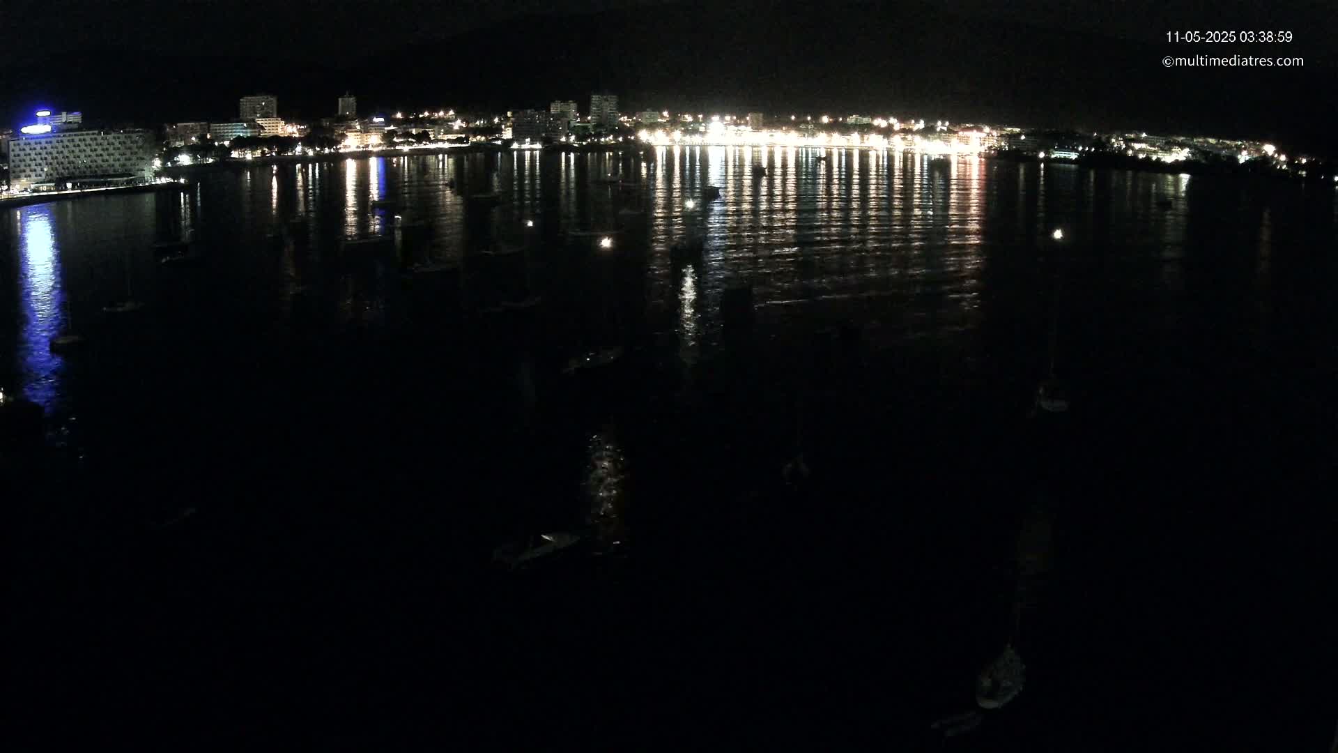 A nighttime view of a city's illuminated waterfront, showing numerous boats moored in the dark water, reflecting the city lights.