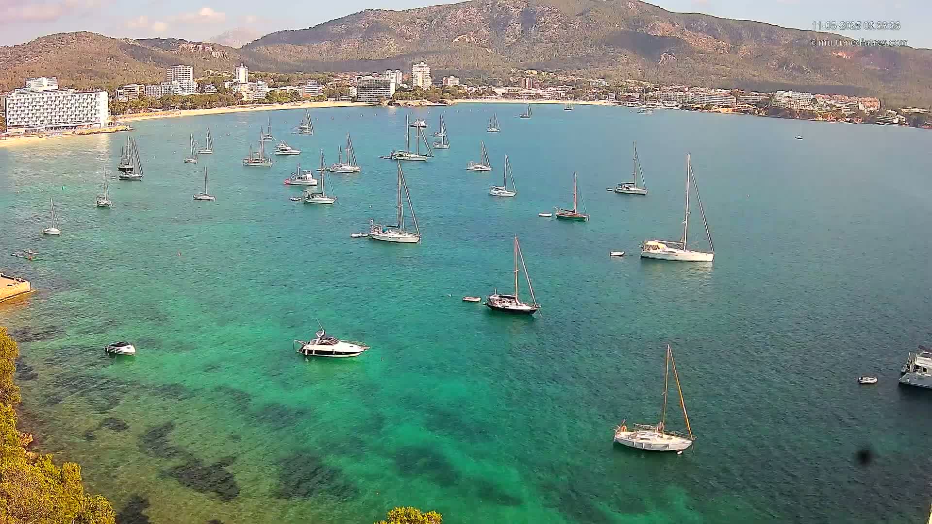 Numerous sailboats and motorboats are anchored in clear, teal water near a beach and buildings nestled against a mountain range under a mostly sunny sky.