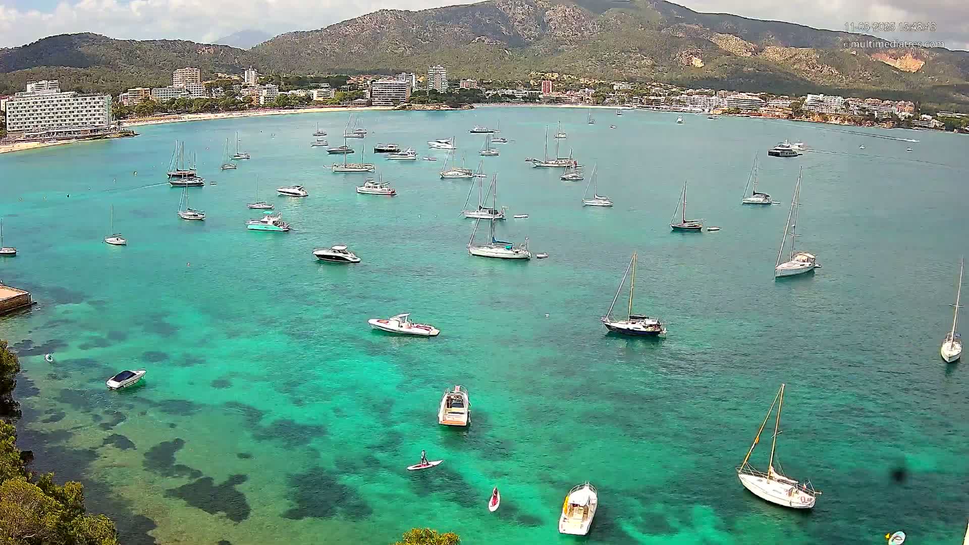 Numerous sailboats and motorboats are anchored in a calm, bright teal bay, with a mountainous coastline and hotels visible in the background under a partly cloudy sky.