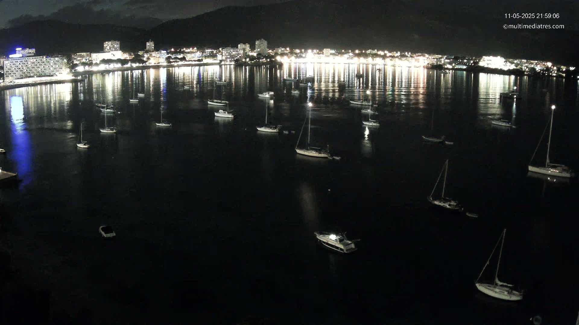 A nighttime view of a calm bay with numerous boats moored, reflecting the lights of a brightly lit city on the shore.