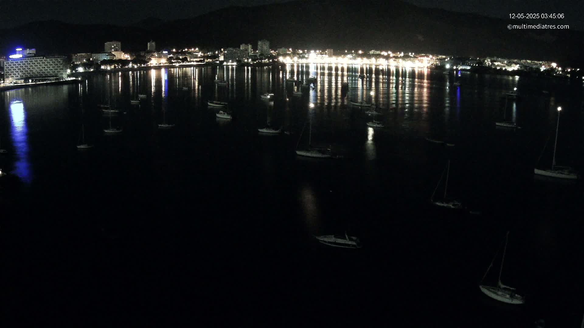 At night, many sailboats are moored in a dark bay in front of a brightly lit town, reflected in the calm water.