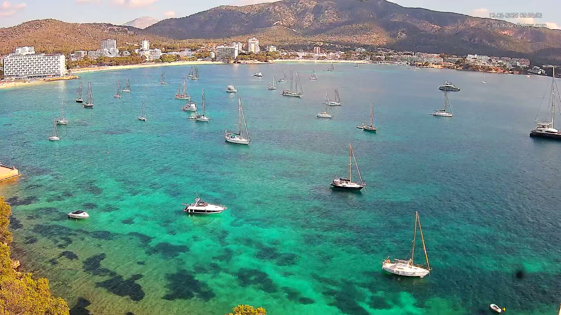 Numerous sailboats and motorboats are anchored in a calm, clear, turquoise bay, with a mountain range and a shoreline of hotels in the background on a sunny day.