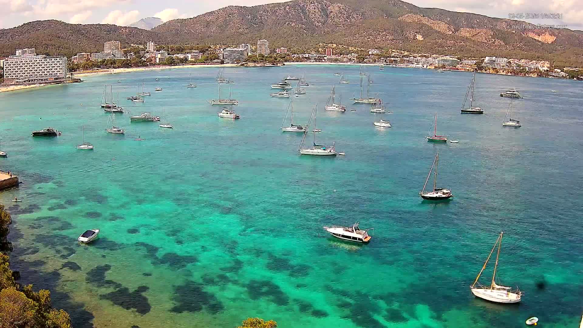 Numerous sailboats and motorboats are anchored in a calm, clear turquoise bay, with a mountainous coastline and buildings in the background under sunny skies.