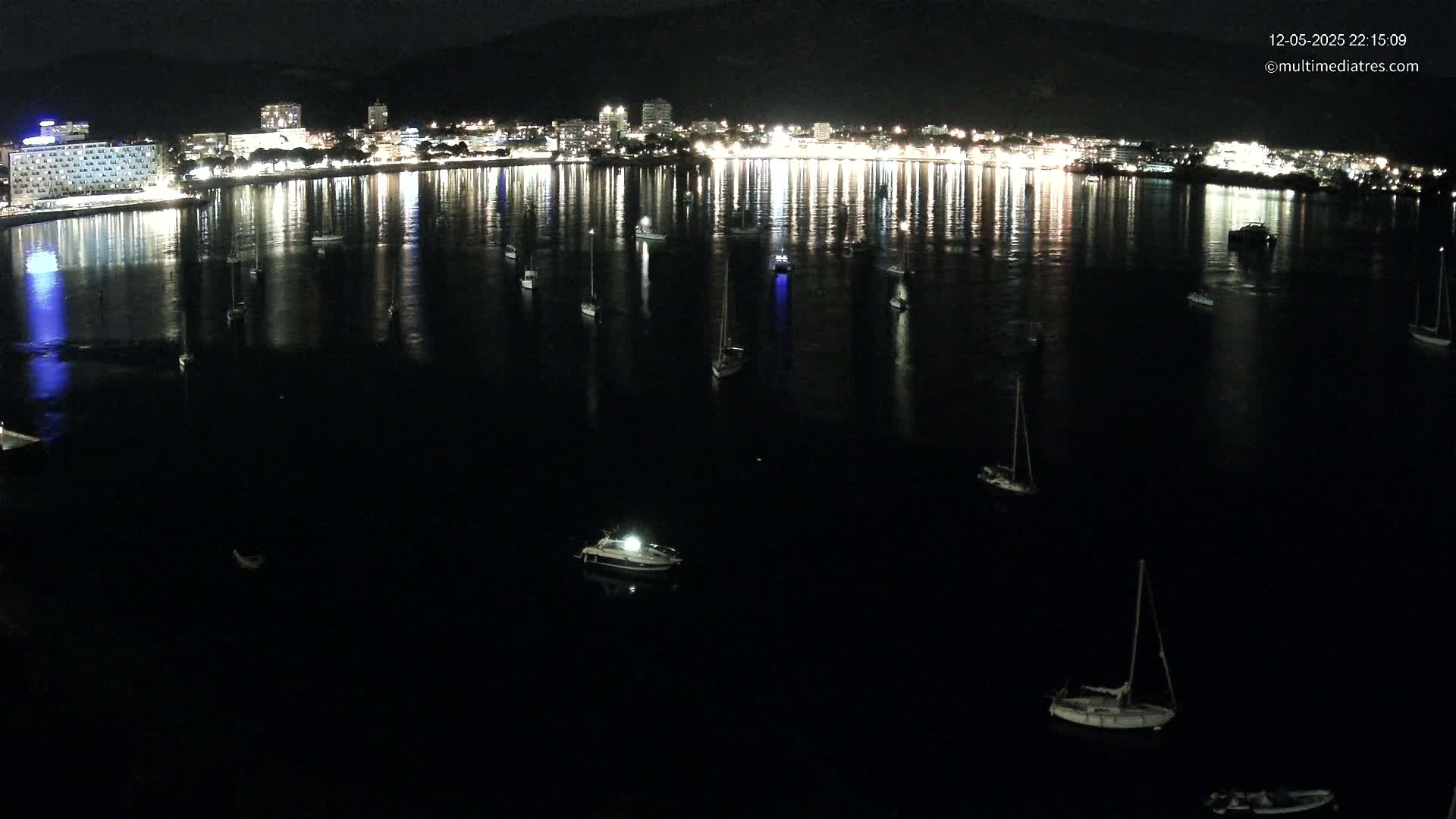 A nighttime view of a calm body of water with numerous boats and a brightly lit city reflected in the water.