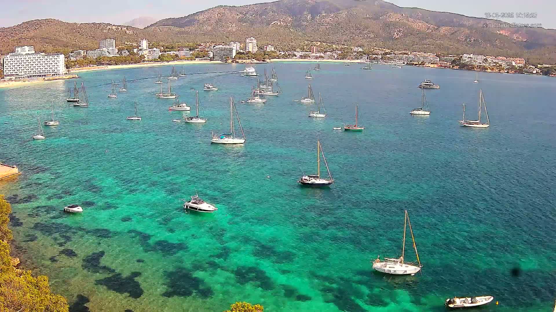 Numerous sailboats and motorboats are anchored in a calm, clear turquoise bay near a shoreline with hotels and a mountainous backdrop under a sunny sky.