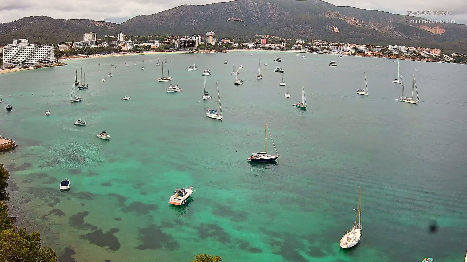 Numerous boats are anchored in a calm, teal bay, with a shoreline of buildings and a mountain range visible in the background under an overcast sky.