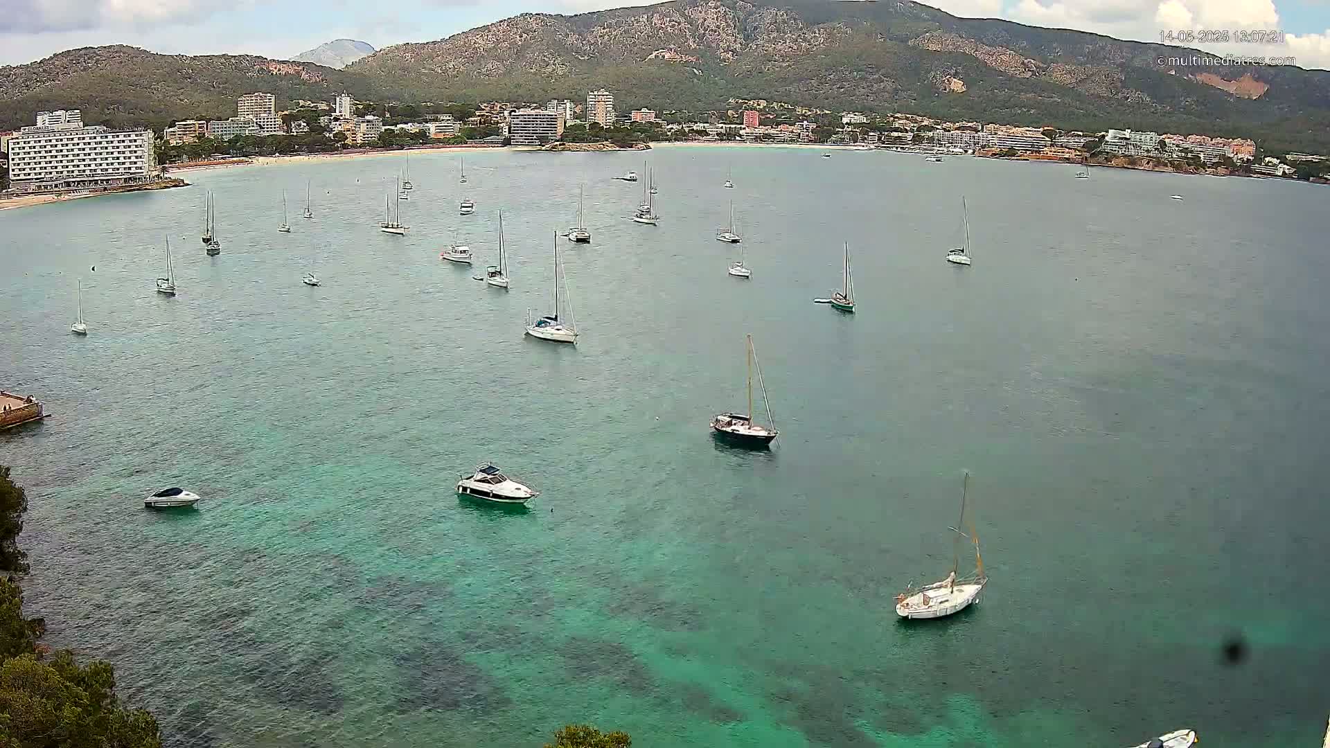Numerous sailboats and motorboats are anchored in a calm, clear turquoise bay near a mountainous coastline with hotels and buildings visible.