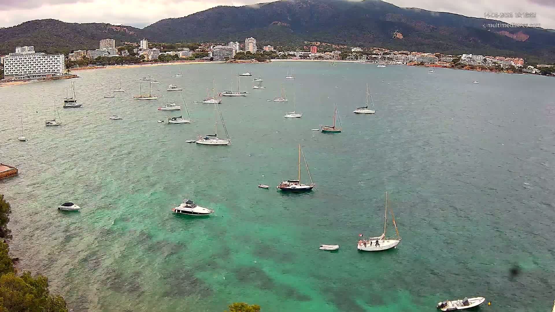Numerous sailboats and motorboats are anchored in a calm, teal bay near a coastline with hotels and mountains under an overcast sky.