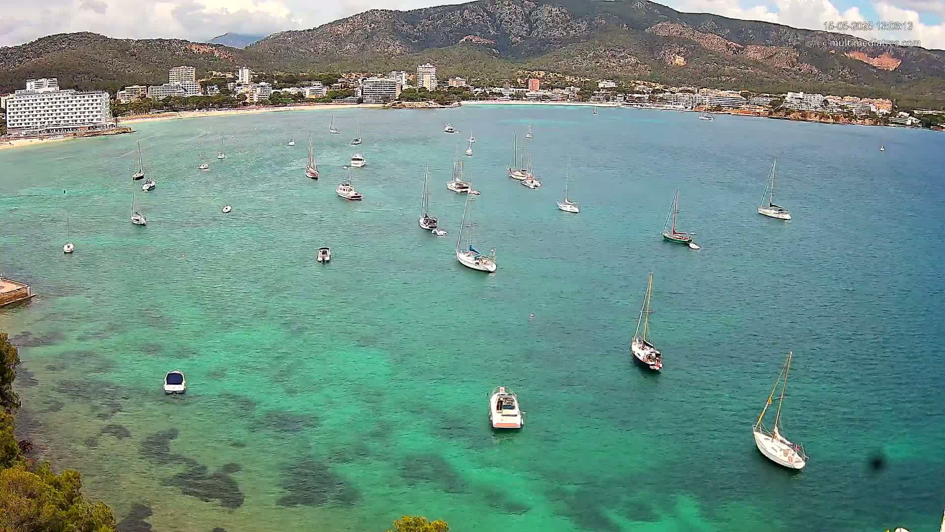 Numerous sailboats and motorboats are anchored in a calm, clear, teal-colored bay, with a mountainous coastline and hotels visible in the background under a partly cloudy sky.