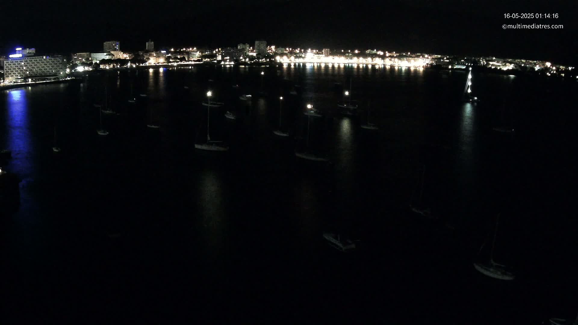 Many small boats are moored in a dark, calm body of water at night, in front of a brightly lit city skyline.