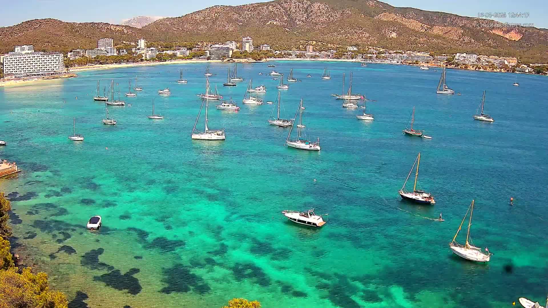 Numerous sailboats are anchored in a calm, clear turquoise bay near a beach and buildings, backed by mountains under a sunny sky.