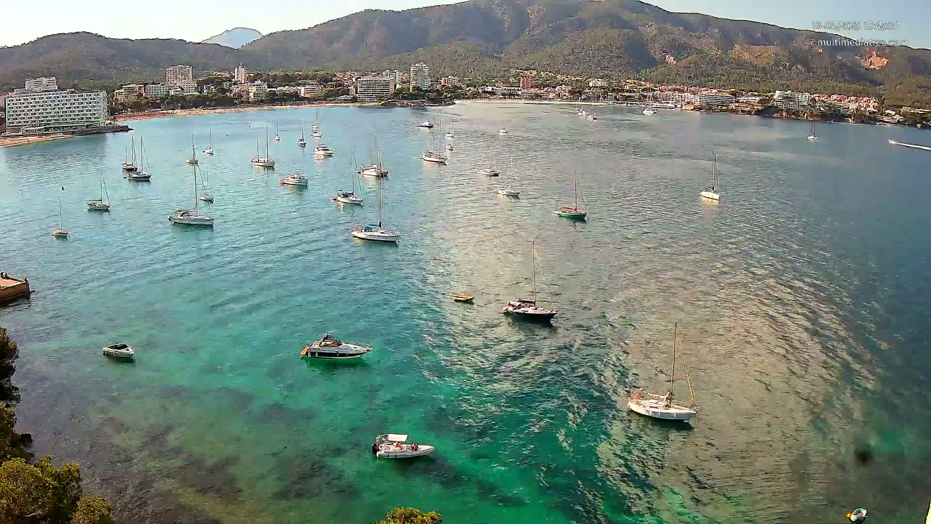 Numerous sailboats and motorboats are anchored in a calm, clear turquoise bay, with a shoreline of hotels and buildings backed by mountains in the distance.