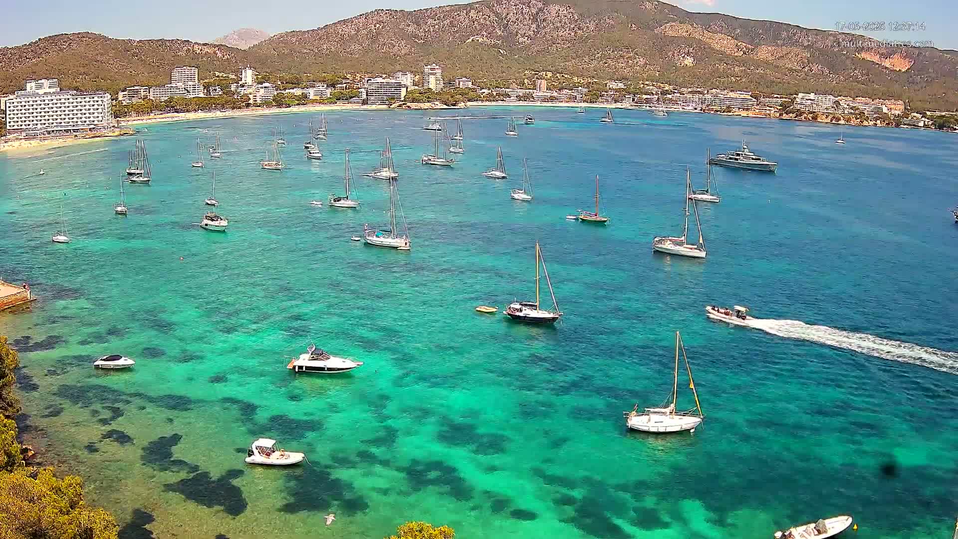 Numerous sailboats and motorboats are anchored in a clear, turquoise bay near a beach and hotels situated against a backdrop of mountains under a sunny sky.