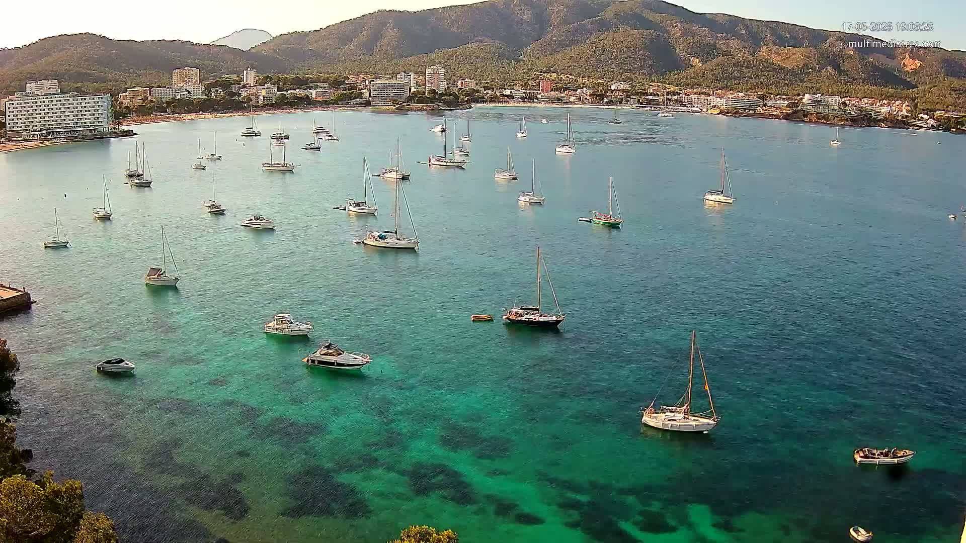 Numerous sailboats and motorboats are anchored in a calm, clear, teal-colored bay, with a coastline of hotels and mountains in the background under a clear sky.