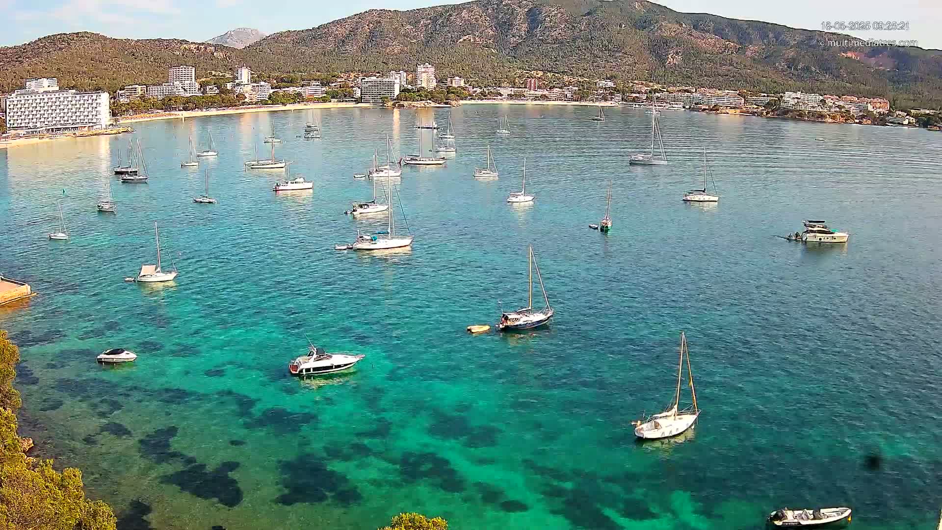 Numerous sailboats and motorboats are anchored in a calm, clear turquoise bay, with a mountainous shoreline and buildings in the background under sunny conditions.