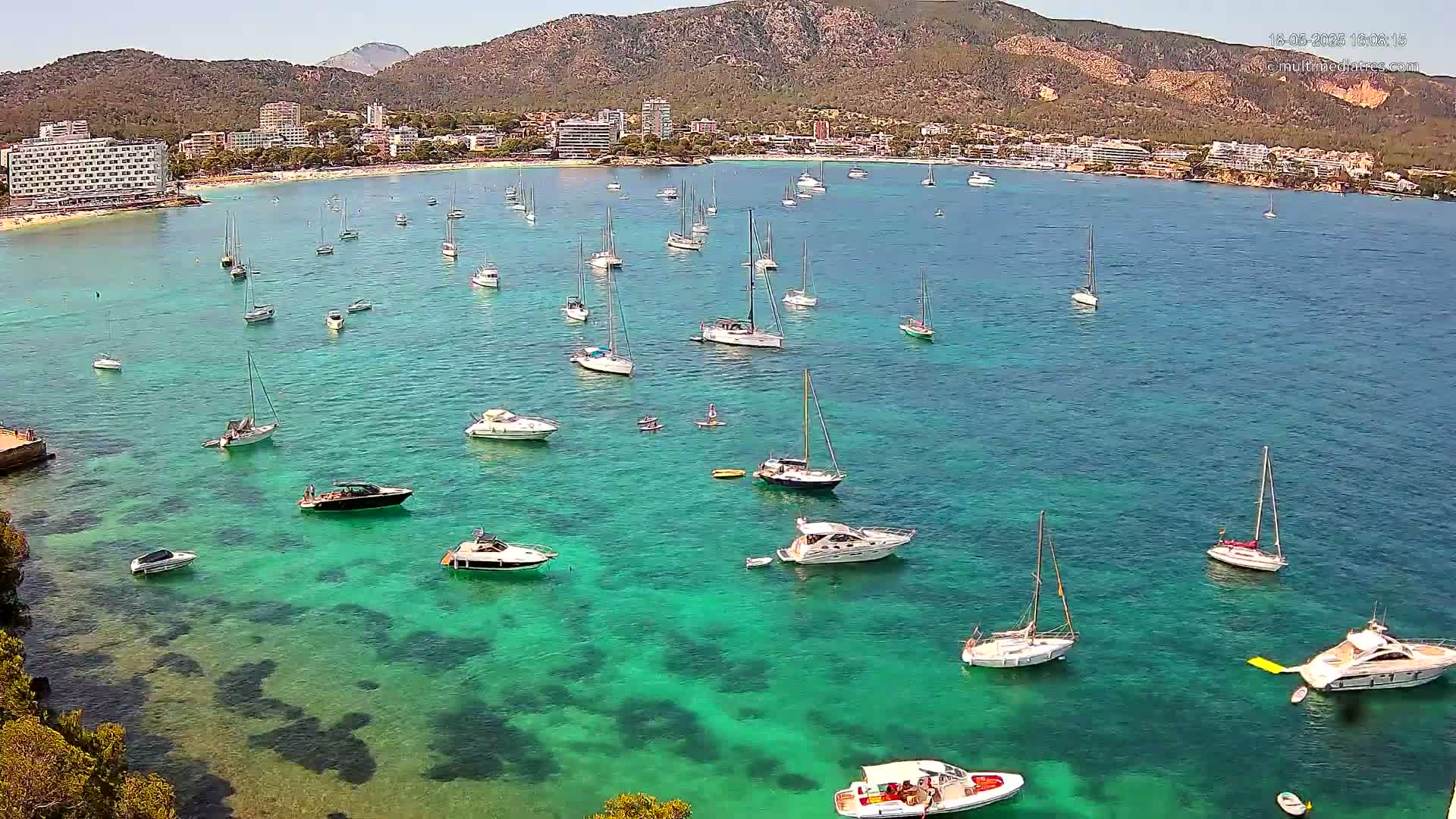 Numerous boats and yachts are anchored in clear, turquoise water near a beach and hotels situated against a mountainous backdrop under sunny skies.