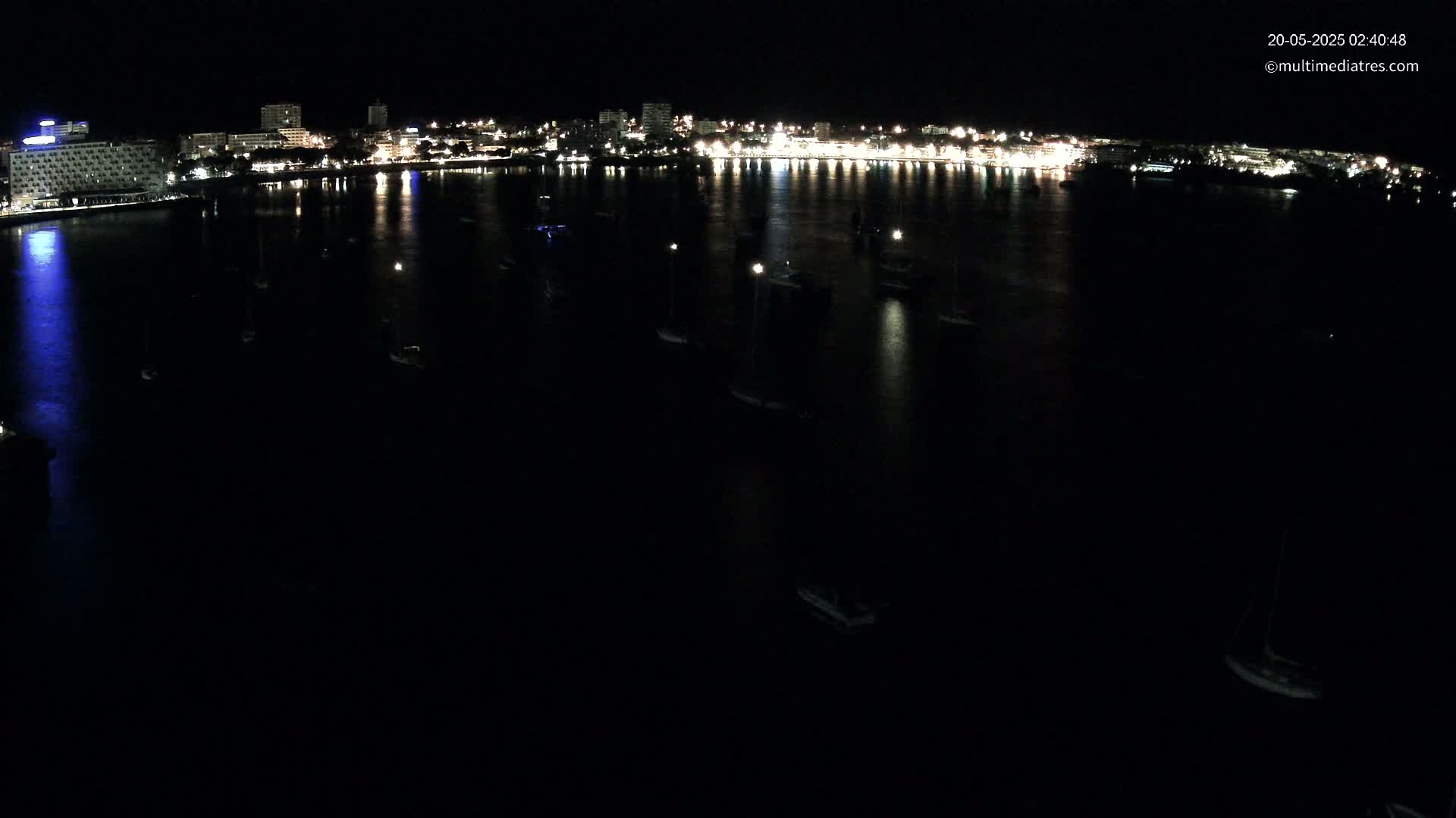 A nighttime aerial view shows a city's illuminated waterfront with numerous boats moored in calm, dark water.