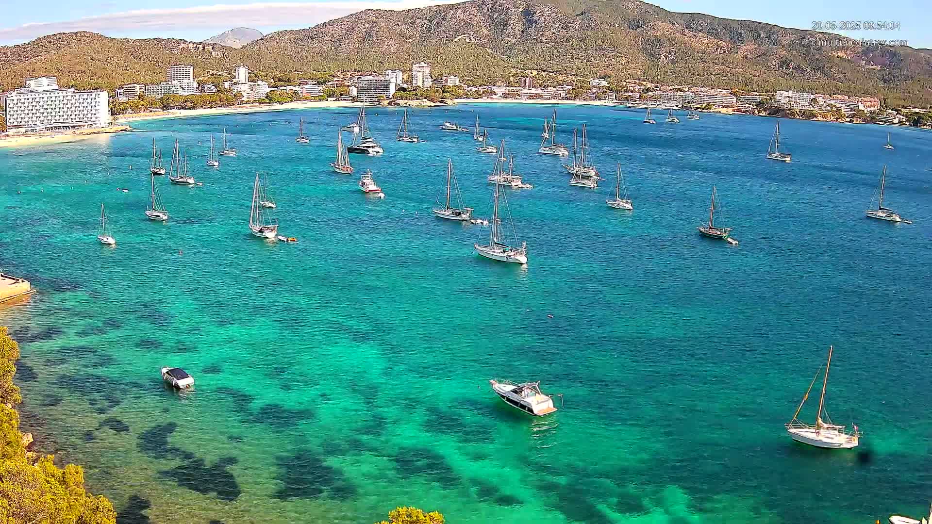 A tranquil bay with clear, turquoise water is dotted with numerous sailboats and motorboats, all anchored near a sandy beach backed by hotels and a mountain range under a sunny sky.