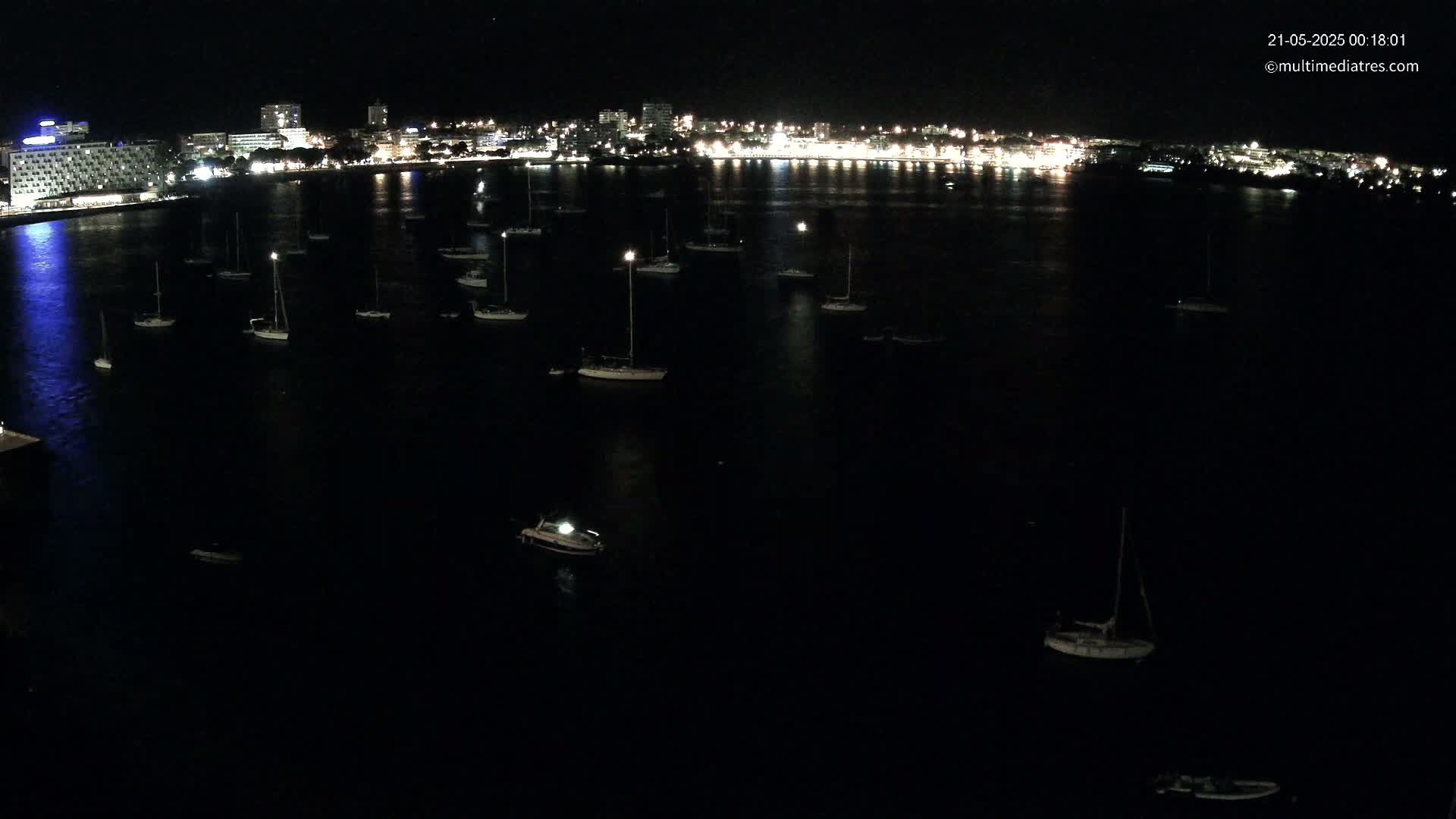 Numerous sailboats are moored in a dark body of water at night, with a brightly lit city in the background.