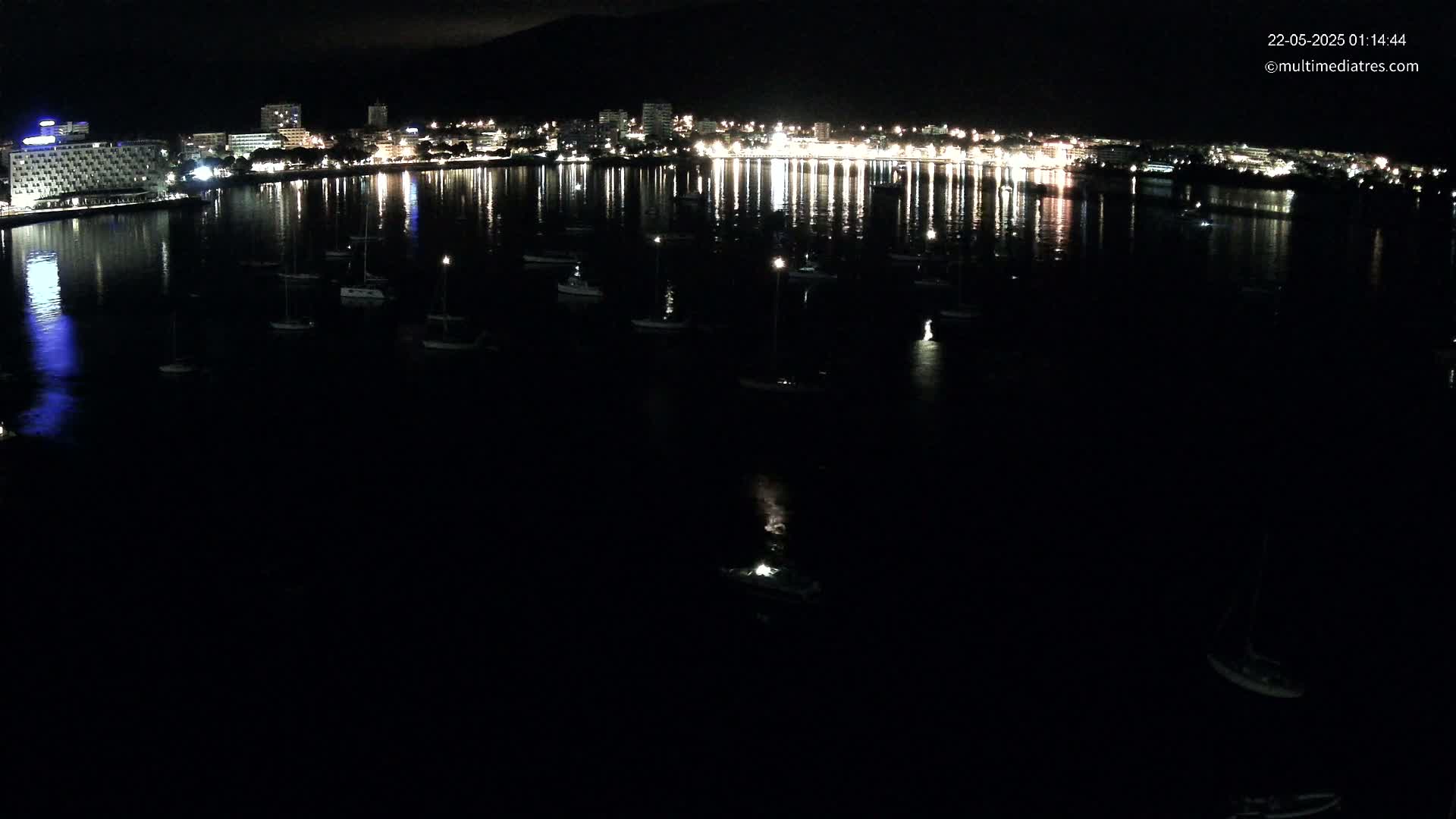 A nighttime view of a calm body of water with numerous boats and a brightly lit city reflected in the water.