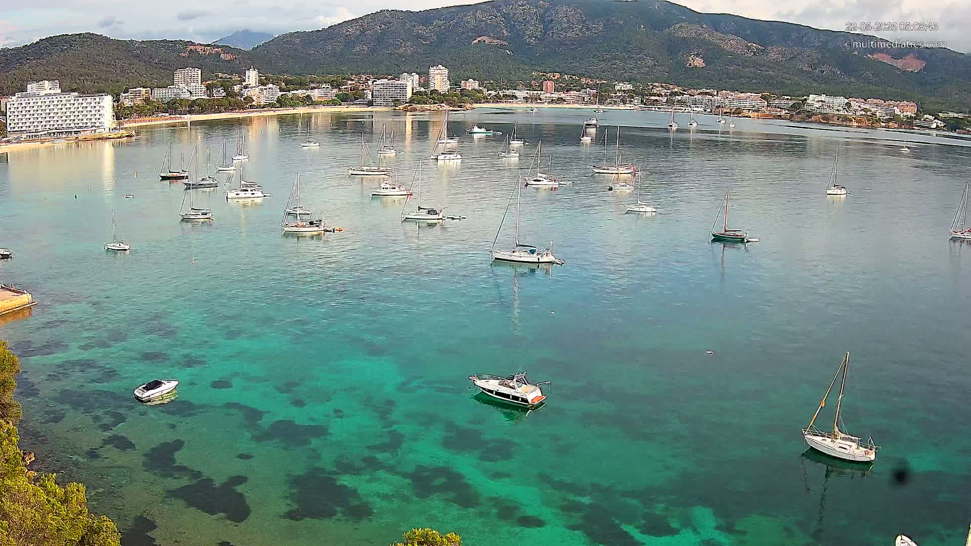 A calm bay with numerous sailboats and motorboats anchored in clear, turquoise water, with a mountain range and coastal buildings visible in the background under a partly cloudy sky.